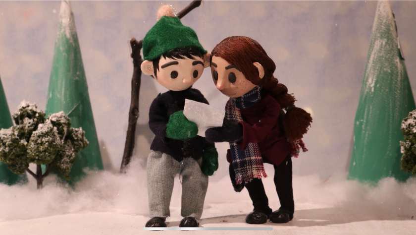 Claymation wintery scene with snow covered trees and a woman and man looking down at a piece of paper. 