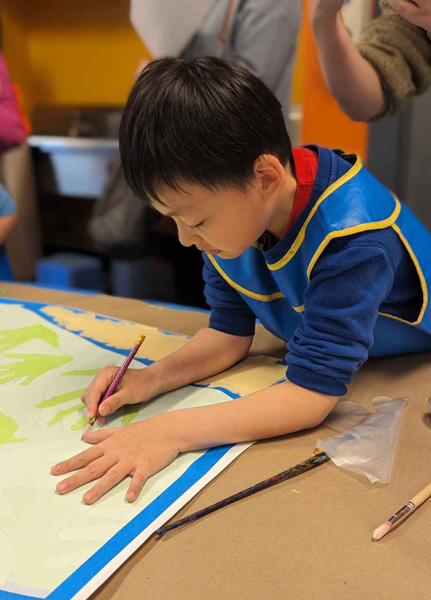 A child sitting at a table, painting on a large piece of paper while leaning over the table. 