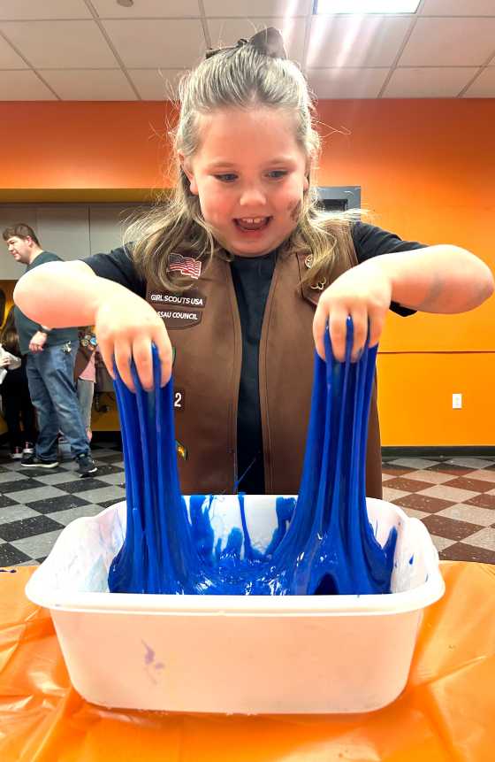 Photograph of a child wearing a brown vest happily pulling blue slime out of a white bin.