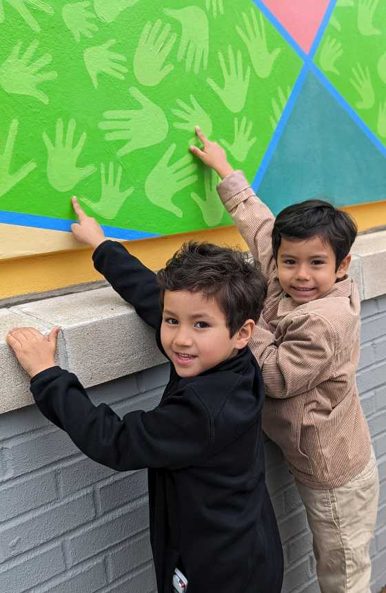 Two children standing in front of a painted mural of hand prints while pointing to the prints. 