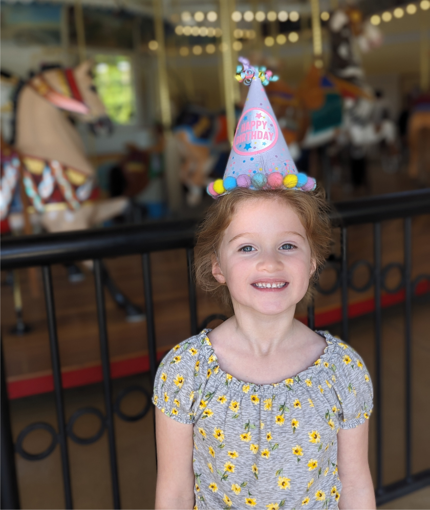 A child smiling and wearing a party hat in the foreground with Nunley's Carousel in the background. 