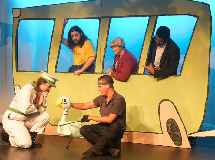 A large cardboard green bus with three actors in the window and the bus driving crouching in front of the bus talking to the Pigeon puppet. 