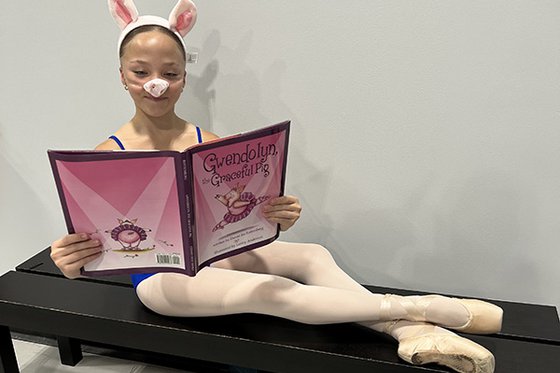 A young woman wearing a ballet leotard and tights, a pig nose and pig ears holding the book Gwendolyn the Graceful Pig while sitting on a bench with their toes pointed.