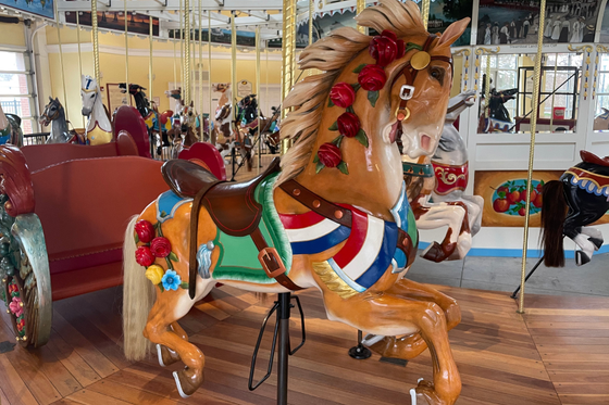 A close-up of brown carousel horse decorated with flowers and a red, white and blue sash with other carousel horses in the background.. 