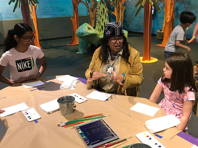 A photograph of an adult talking to two children while they sit at a table doing arts and crafts.