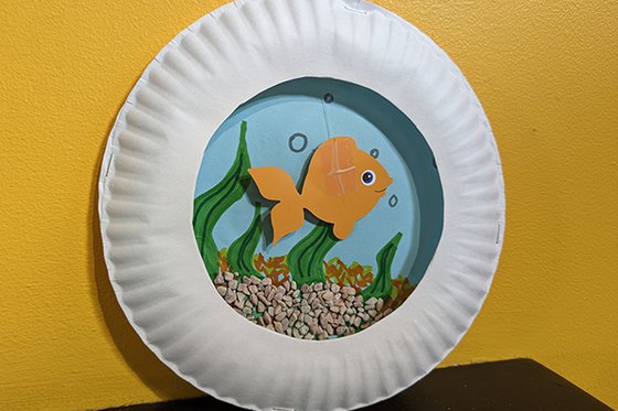 An orange paper goldfish in a paper plate 'bowl' against a blue background with a rock bottom. 