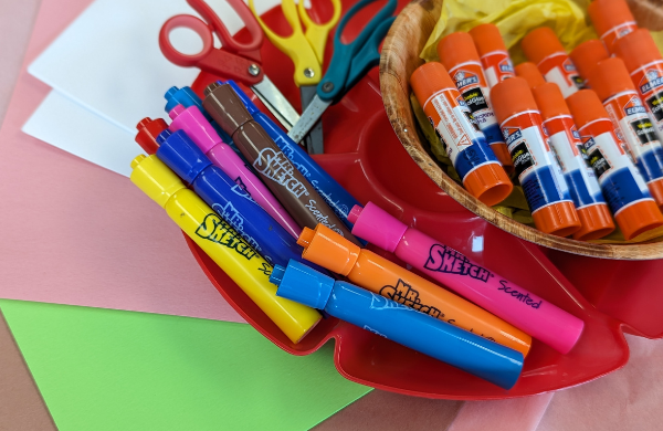 A tray holding glue sticks, scissors and markers on top of colored paper. 