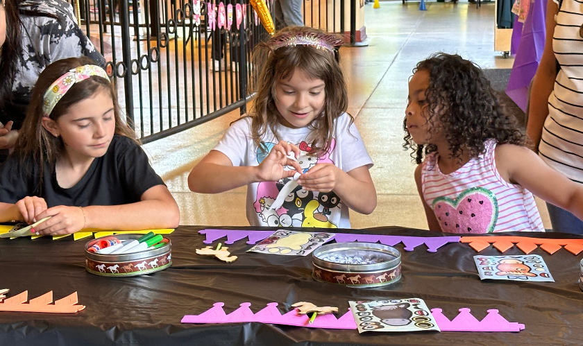 Three children sitting at a table making a carousel themed craft. 