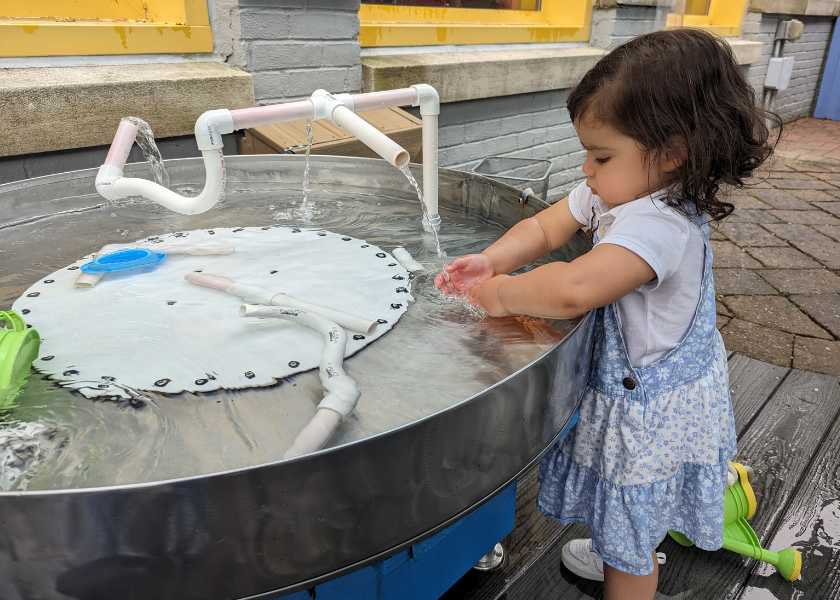 A child standing at a metal water table with thin PVC pipes pouring water onto her hands. 