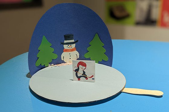 A white circular piece of cardboard mimicking a ice rink with a picture of an ice skating penguin holding a hockey stick, and a blue circular paper background decorated with a snowperson, and trees.  