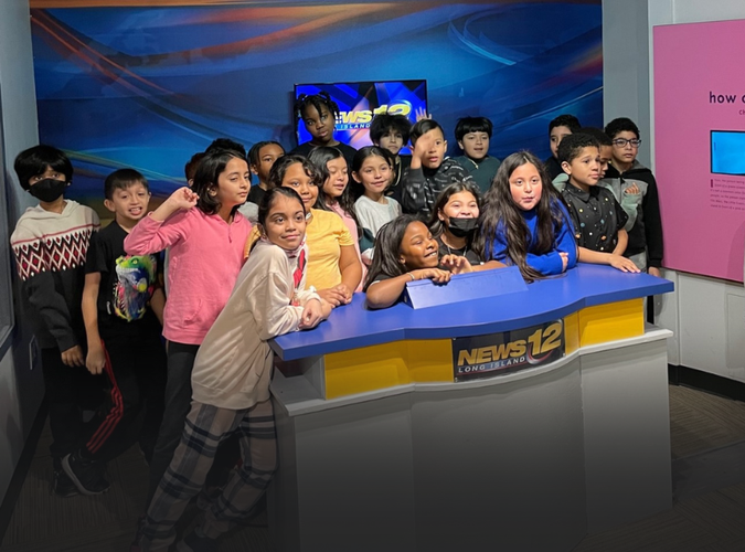 Photo of a class of students gathered around the News 12 Station table. 
