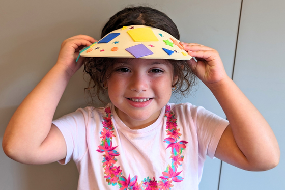 A child smiling at the camera while holding a decorated white visor hat on their head. 