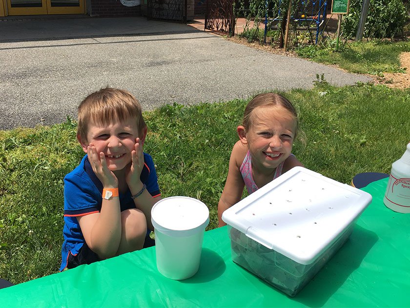 Two children outside at a green table smiling at the camera. There is a white box on the table.