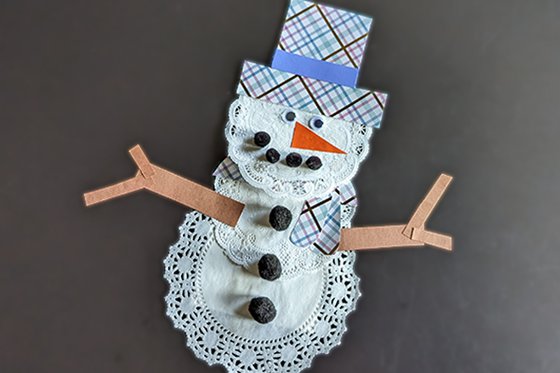 A snowperson made from white paper doilies, with brown arms, a plaid hat, and googly eyes. 