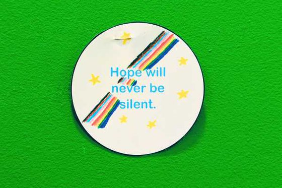 A white circular pin with centered text "Hope will never be silent" with a rainbow going through the text and stars surrounding the text. 