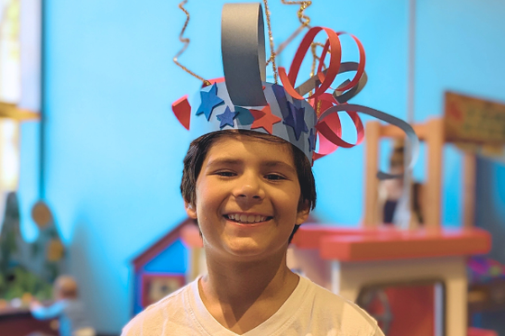A child smiling at the camera while wearing a paper headband decorated with blue and red stars and stripes. 
