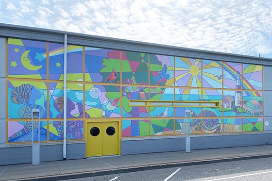 The West side of LICM building featuring a large mural paited in a quilted pattern with pastel colors featuring a sun, tree, island landscape, turtle, people, and birds. 